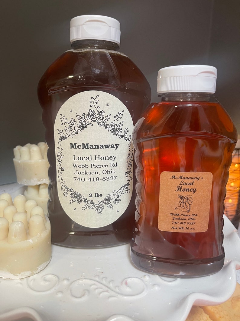 May include: Two bottles of McManaway's Local Honey with white caps. The larger bottle is labeled "2 lbs" and the smaller bottle is labeled "Net Wt. 16 oz." Both bottles have labels with the address and phone number.