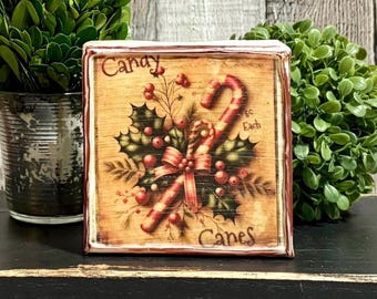 May include: A square wooden sign with a distressed light brown wood grain background. The image features a red and white striped candy cane, green holly leaves, red berries, and a red ribbon. The text reads Candy Canes 5c Each.