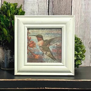 May include: A square, off-white wooden picture frame with a distressed finish. The frame contains a print of a hummingbird in flight near a flower, set against a background of horizontal blue and brown stripes with a weathered, textured appearance.