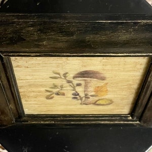 May include: A framed print of a mushroom with a brown cap and white stem, surrounded by leaves and berries. The frame is black with a distressed finish.