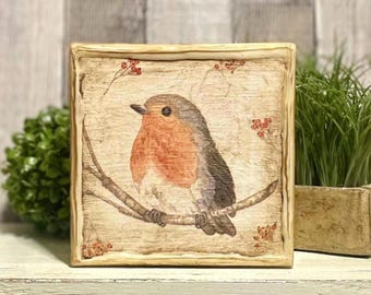 May include: A square wooden plaque featuring a detailed illustration of a robin perched on a branch. The bird has a red breast, gray wings, and is set against a textured, light-brown background. Small red berries and twigs are also depicted.