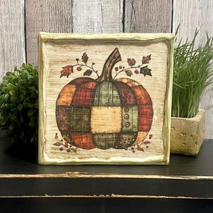May include: Square wooden sign featuring a patchwork pumpkin design. The pumpkin is composed of various fabric patterns in shades of orange, green, and brown. The sign has a distressed, rustic look, with a light green frame and decorative leaves.