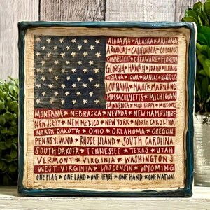 May include: A square, rustic-style plaque featuring the American flag design. The flag's blue field has white stars, and the red stripes are replaced with the names of U.S. states in a handwritten font. The border is dark blue.
