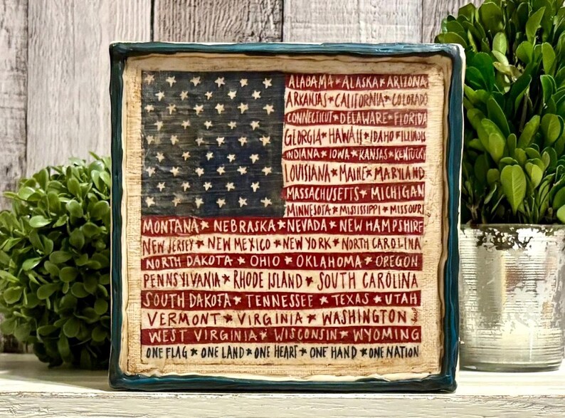 May include: A square, rustic-style plaque featuring the American flag design. The flag's blue field has white stars, and the red stripes are replaced with the names of U.S. states in a handwritten font. The border is dark blue.