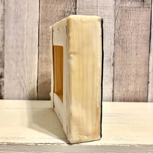 May include: A rectangular, off-white painted canvas with a dark blue edge. The canvas has a rectangular cutout in the center, revealing a tan interior. The canvas is placed on a white, distressed wooden surface.
