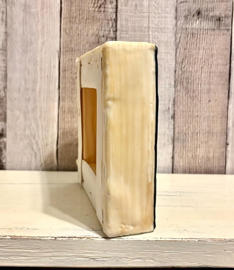May include: A rectangular, off-white painted canvas with a dark blue edge. The canvas has a rectangular cutout in the center, revealing a tan interior. The canvas is placed on a white, distressed wooden surface.