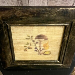 May include: A framed print of a mushroom with leaves and berries on a beige background. The frame is black with a distressed finish.