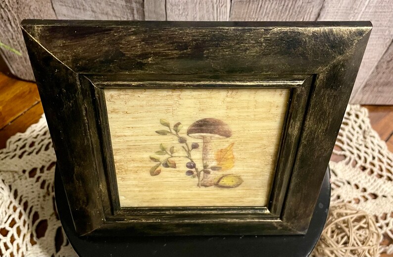 May include: A framed print of a mushroom with leaves and berries on a beige background. The frame is black with a distressed finish.