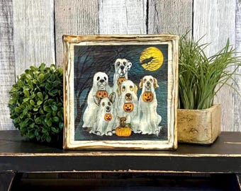 Puede incluir: Un letrero de madera cuadrado con una ilustración impresa de cinco perros disfrazados de fantasmas, cada uno sosteniendo un pequeño cubo de calabaza naranja. La escena incluye una luna llena amarilla, un cuervo negro y árboles oscuros.