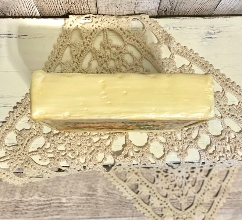 May include: A rectangular bar of ivory-colored soap with a floral design on the bottom. The soap is sitting on a white crocheted doily.