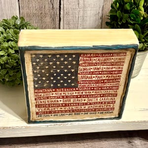May include: A rectangular wooden block featuring a painted American flag design. The flag includes the stars and stripes, with the names of the states listed below. The block has a distressed, vintage look, with the text "ONE FLAG ONE LAND ONE HEART ONE HAND ONE NATION".