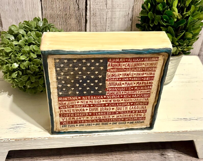 May include: A rectangular wooden block featuring a painted American flag design. The flag includes the stars and stripes, with the names of the states listed below. The block has a distressed, vintage look, with the text "ONE FLAG ONE LAND ONE HEART ONE HAND ONE NATION".