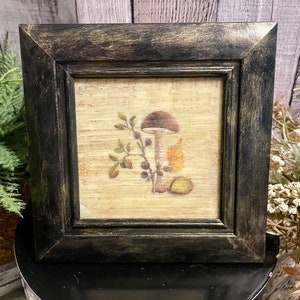 May include: A black framed picture with a beige background featuring a mushroom with brown cap and white stem, surrounded by leaves and berries.