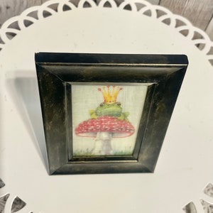 May include: A framed illustration of a green frog wearing a gold crown sitting on a red and white mushroom.