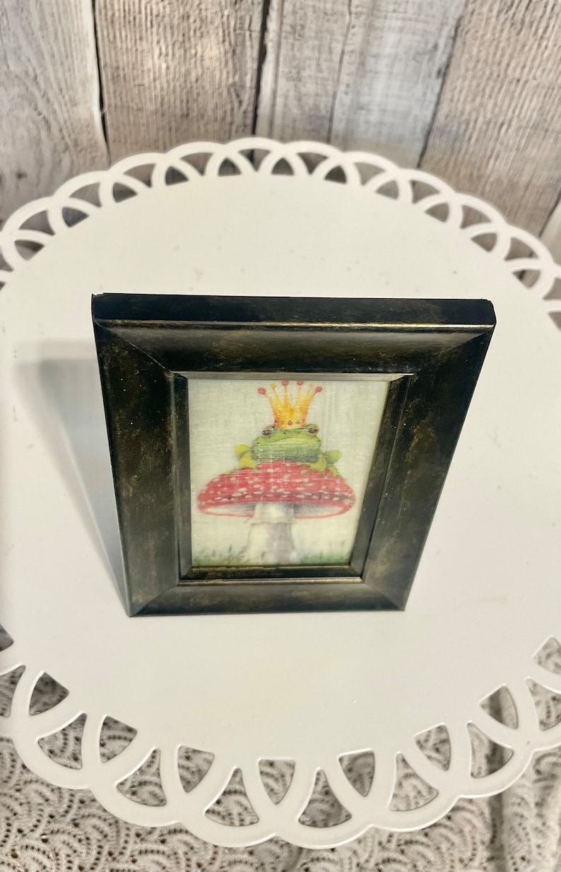 May include: A framed illustration of a green frog wearing a gold crown sitting on a red and white mushroom.
