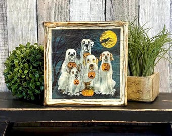 May include: A square wooden sign featuring a printed illustration of five dogs dressed as ghosts, each holding a small orange jack-o'-lantern bucket. The scene is set at night under a full yellow moon with a dark tree silhouette in the background.