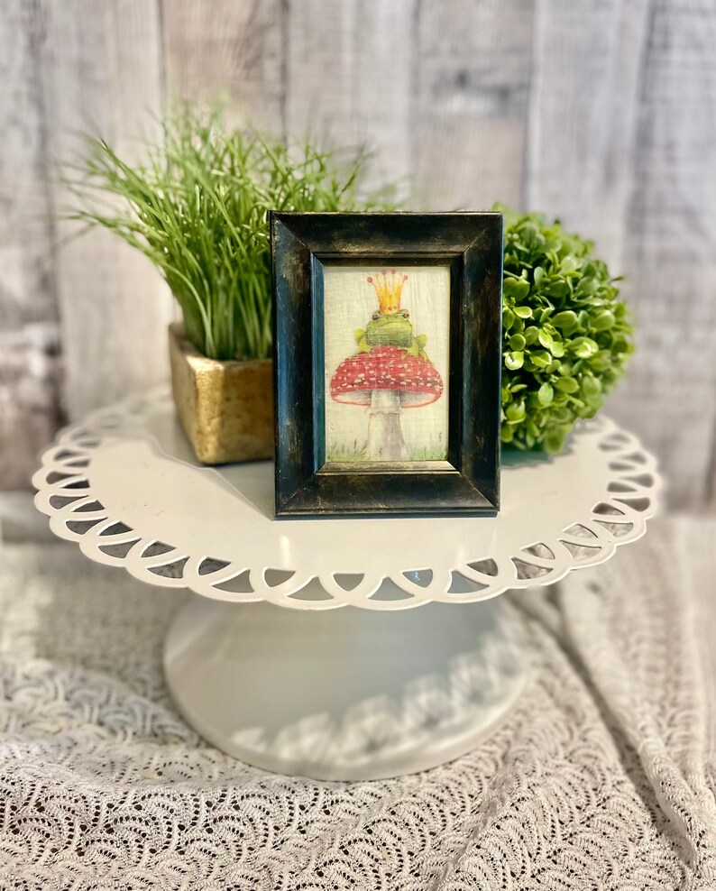 May include: A framed illustration of a green frog wearing a gold crown sitting on a red and white mushroom. The frame is black with a gold inner border.