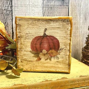 May include: A square wooden block with a painted pumpkin design. The pumpkin is a deep red color with a green stem and is surrounded by fall leaves. The block has a distressed, rustic look with a light brown background and a gold border.