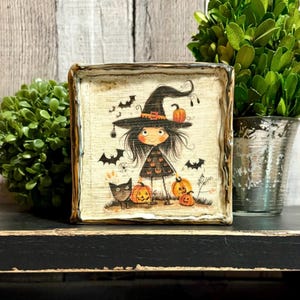 May include: A square wooden sign featuring a whimsical illustration of a witch with long black hair wearing a pointed hat, surrounded by orange pumpkins, black bats, and a small black cat on a cream-colored background with a distressed edge.