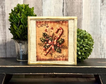 May include: A square wooden sign with a distressed cream frame featuring a vintage-style illustration of a candy cane, holly leaves, and red berries. The text Candy Canes is printed in a dark brown script font on a warm, wood-toned background.