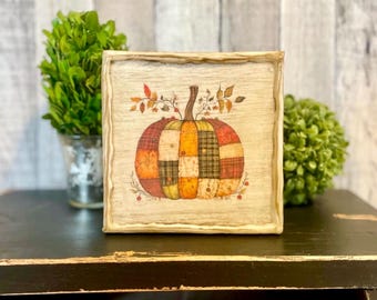 May include: A square wooden sign featuring a central illustration of a pumpkin composed of various patchwork fabric patterns in shades of orange, yellow, green, and brown, topped with small leafy branches and a stem on a light wood background.