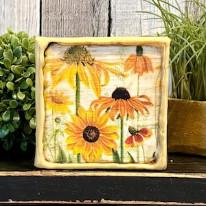 May include: A square wooden art piece featuring a floral design of yellow and orange coneflowers. The artwork is framed in a light yellow border and set against a rustic wooden background. The piece is displayed on a black surface.