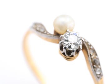 May include: A close-up of a gold ring with a pearl and a diamond. The ring has a curved design with a pearl at the top and a diamond below it. The band is gold with a silver-colored accent.