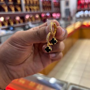 May include: A pair of gold hoop earrings featuring small clear stones along the outer edge and a dangling black four-leaf clover charm. The earrings are constructed with a hinged closure mechanism and a polished gold finish, showcasing a delicate design.