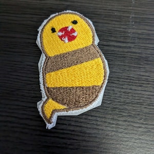 May include: Embroidered patch featuring a cartoon bird with a yellow head and brown striped body. The bird has a red and white open mouth and small black eyes. The patch is outlined in white and has a paper backing.