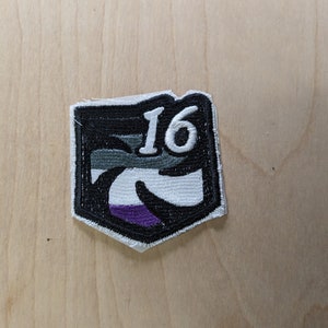 May include: Embroidered patch with a black border and a shield shape. The patch features the number "16" in white, set against a design with black, gray, white, and purple colors. The patch is on a light brown wooden surface.