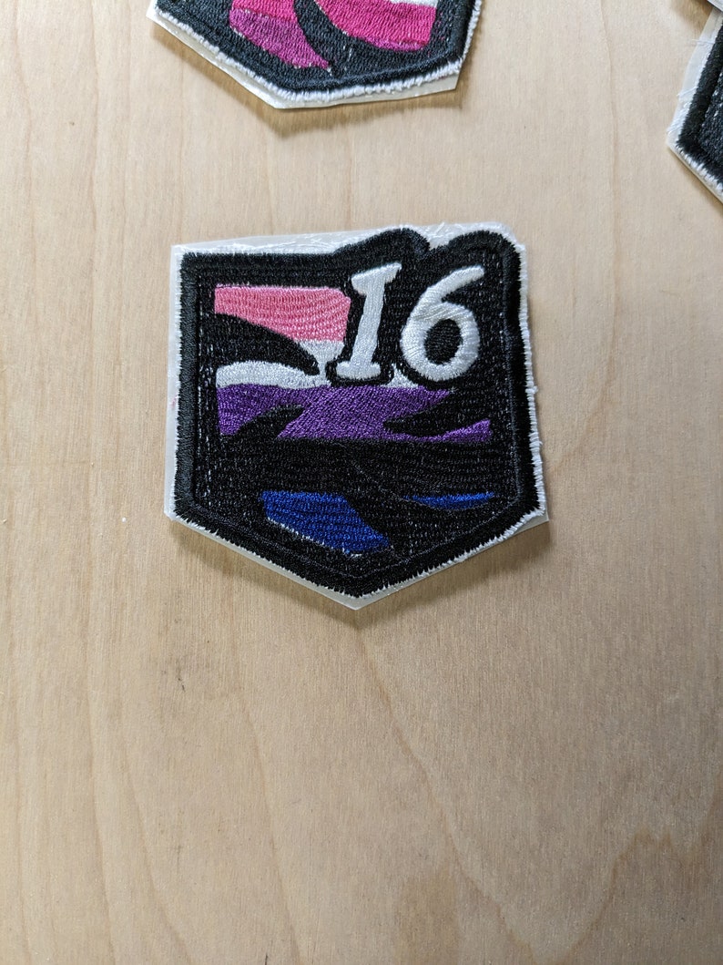 Pu&ograve; includere: Una toppa ricamata in bianco e nero con un design a strisce viola, bianche e rosa. Il numero "16" &egrave; ricamato in bianco sulla toppa.