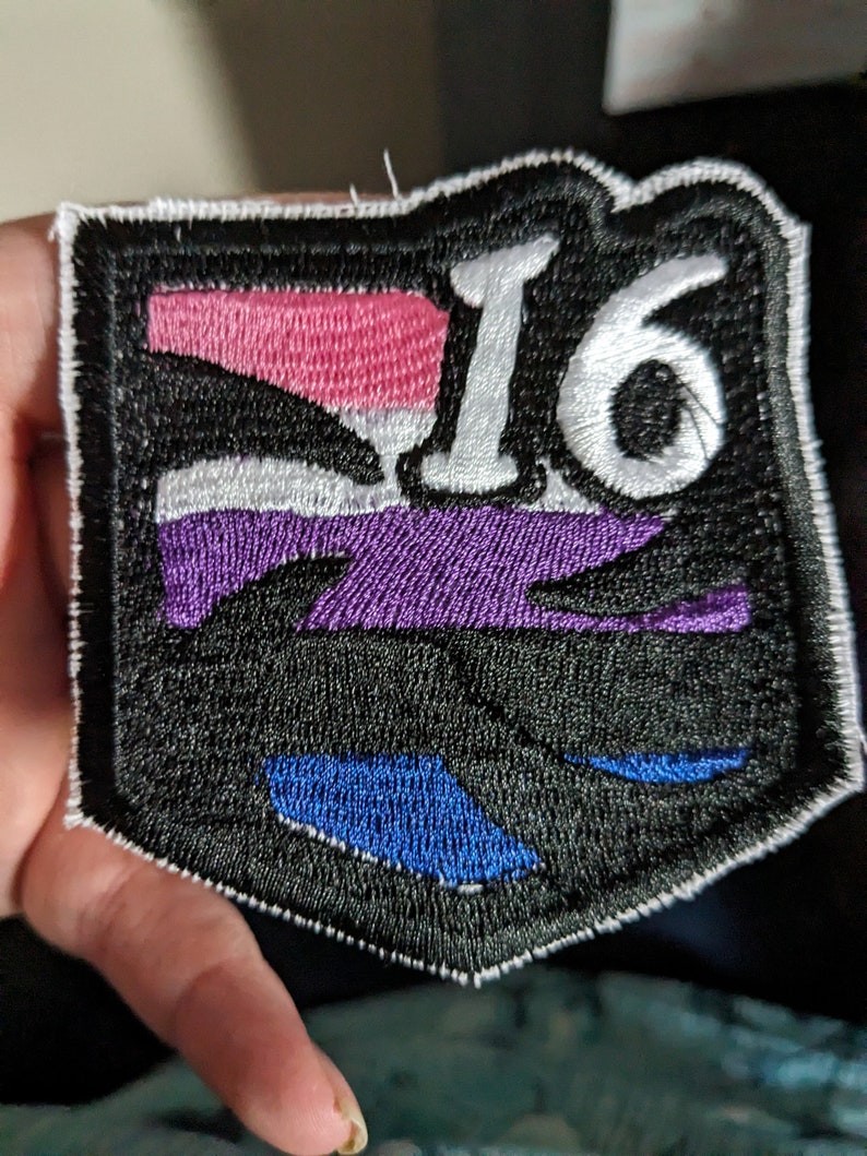 Pu&ograve; includere: Toppa ricamata nera con un "16" bianco al centro. La toppa presenta un design a strisce viola, bianca e rosa con accenti blu.