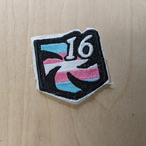 以下が含まれることがあります： 白黒の刺繍パッチで、トランスジェンダーのプライドフラッグのデザインと白い数字「16」が描かれています。