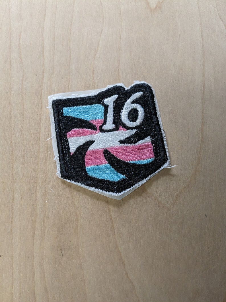 以下が含まれることがあります： 白黒の刺繍パッチで、トランスジェンダーのプライドフラッグのデザインと白い数字「16」が描かれています。
