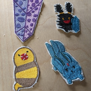 May include: Four embroidered patches on a wooden surface. The patches include a purple crystal, a black creature with a shield, a yellow and brown bird, and a blue rabbit. Each patch has a white border.
