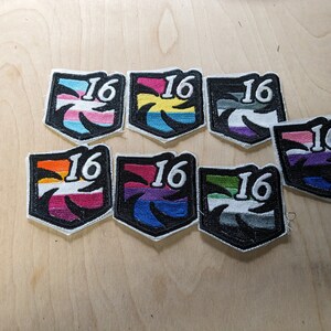 Pu&ograve; includere: Otto toppe ricamate con un designo in bianco e nero di una palma e il numero 16. Ogni toppa ha un diverso motivo a strisce colorate, tra cui rosa, giallo, viola, grigio, arancione, blu, verde e bianco.