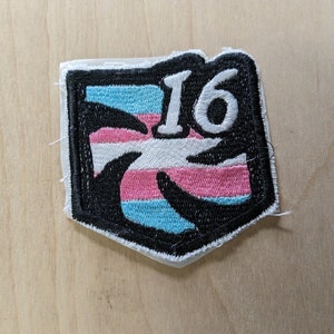 以下が含まれることがあります： 黒と白の刺繍パッチで、トランスジェンダーのプライドフラッグのデザインと白い数字「16」が描かれています。