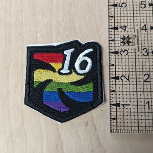以下が含まれることがあります： 虹色のデザインと白い数字「16」が刺繍された黒と白のワッペン。ワッペンは約5cm幅、5cm高さです。