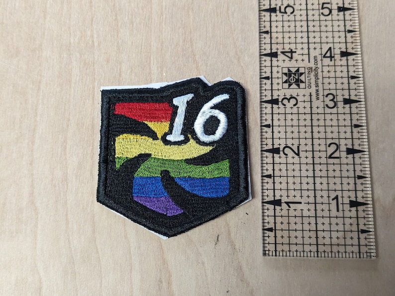 以下が含まれることがあります： 虹色のデザインと白い数字「16」が刺繍された黒と白のワッペン。ワッペンは約5cm幅、5cm高さです。