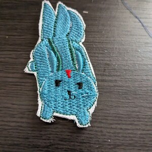 May include: A blue embroidered bunny patch with a red triangle on its forehead. The bunny has black eyes and a white outline.