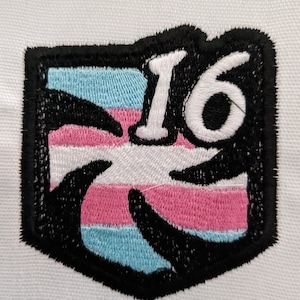 May include: A black and white embroidered patch with a transgender pride flag design and the number "16" in white.