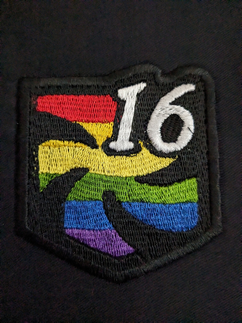 Puede incluir: Un parche bordado con un dise&ntilde;o de arco&iacute;ris y el n&uacute;mero 16 en letras blancas.