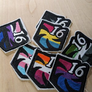 以下が含まれることがあります： ヤシの木のデザインと数字の16が刺繍された8つのパッチのセット。各パッチは、トランスジェンダーのプライドフラッグの異なる色の組み合わせを特徴としています。