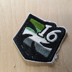 May include: Black and white embroidered patch with a green and white design. The patch has the number "16" in white on it.