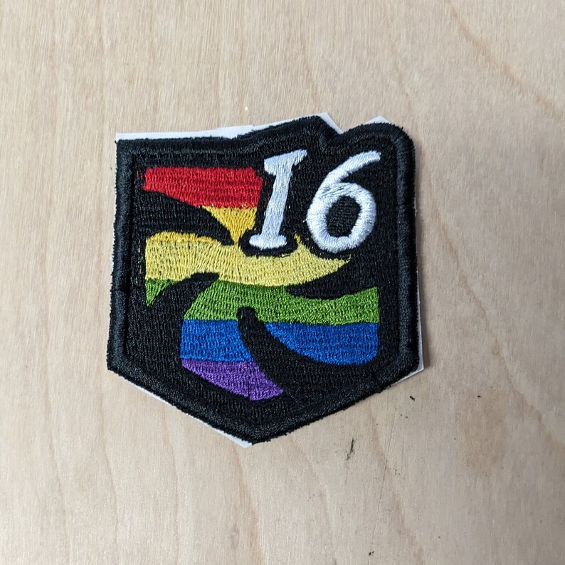 Puede incluir: Un parche bordado negro y arco&iacute;ris con el n&uacute;mero "16" en blanco.