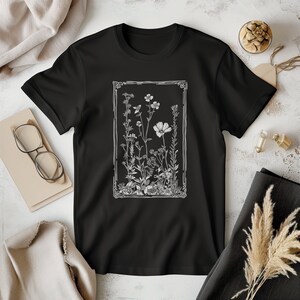 Może przedstawiać: Czarny T-shirt z białym nadrukiem vintage botanicznej ilustracji kwiatów i liści w ozdobnej ramie.