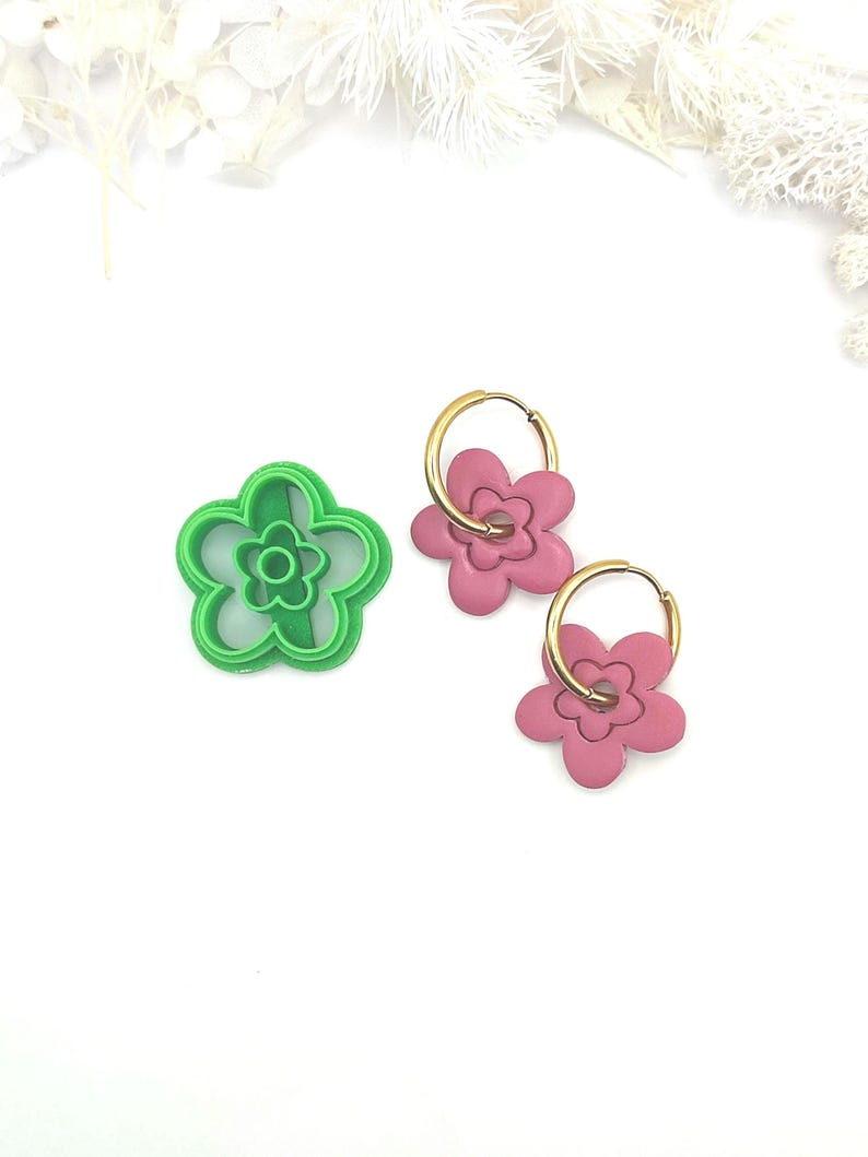 May include: A green flower-shaped cookie cutter and two pink flower earrings with gold hoops are displayed on a white surface. The earrings and cutter have a retro, floral design. The earrings are a dangle style.