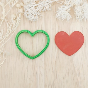 Op de afbeelding: Een groene hartvormige koekjesuitsteker naast een rode hartvormige uitsparing. De koekjesuitsteker heeft een dubbele omtrek. De achtergrond is een licht houten oppervlak met witte decoratieve elementen.
