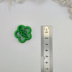 May include: A bright green, flower-shaped cookie cutter. The cutter has a five-petal design with a smaller flower shape in the center. A ruler is placed next to the cutter for size reference.