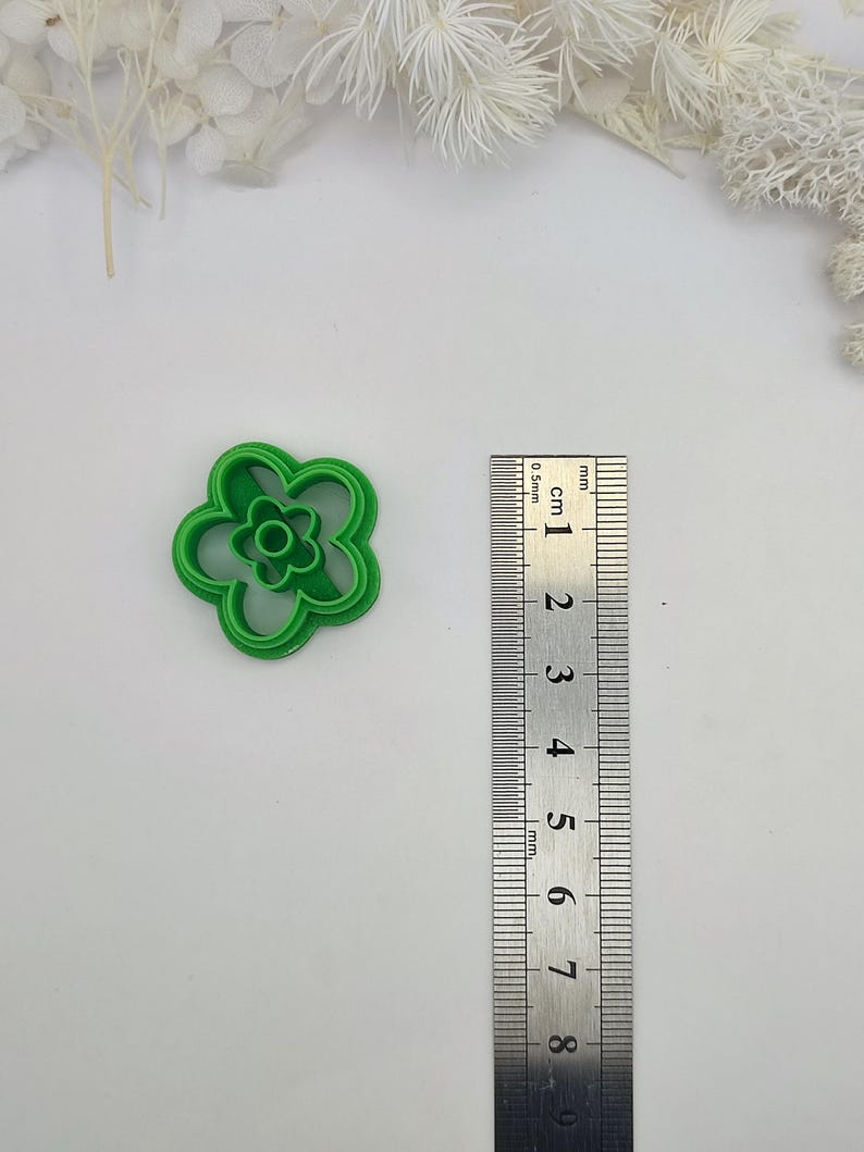 May include: A bright green, flower-shaped cookie cutter. The cutter has a five-petal design with a smaller flower shape in the center. A ruler is placed next to the cutter for size reference.